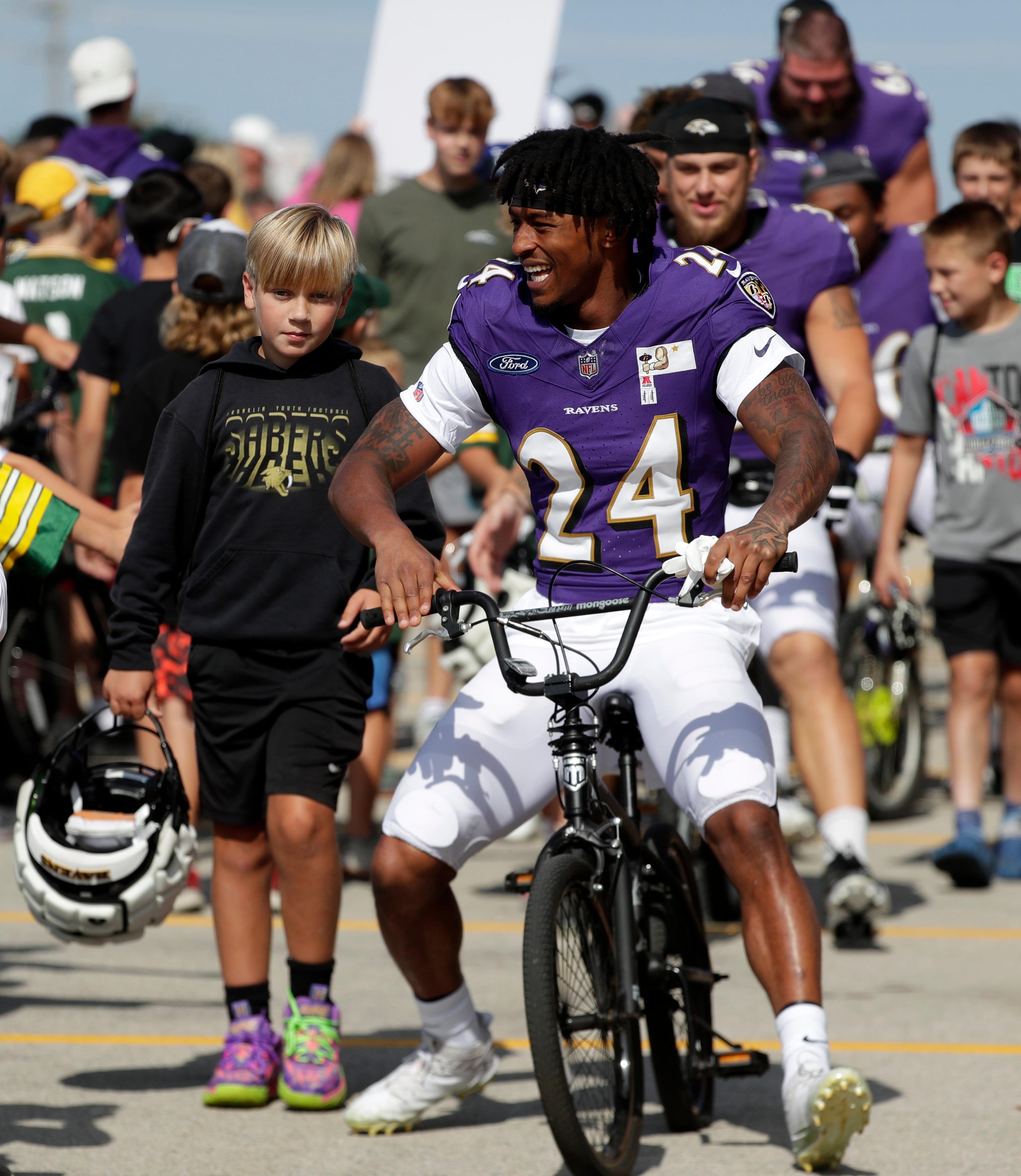 Baltimore Ravens cornerback Damarion Williams (24) rides a young fan's bicycle to a joint practice with the Green Bay Packers on Aug. 22, 2024 in Green Bay, Wis.