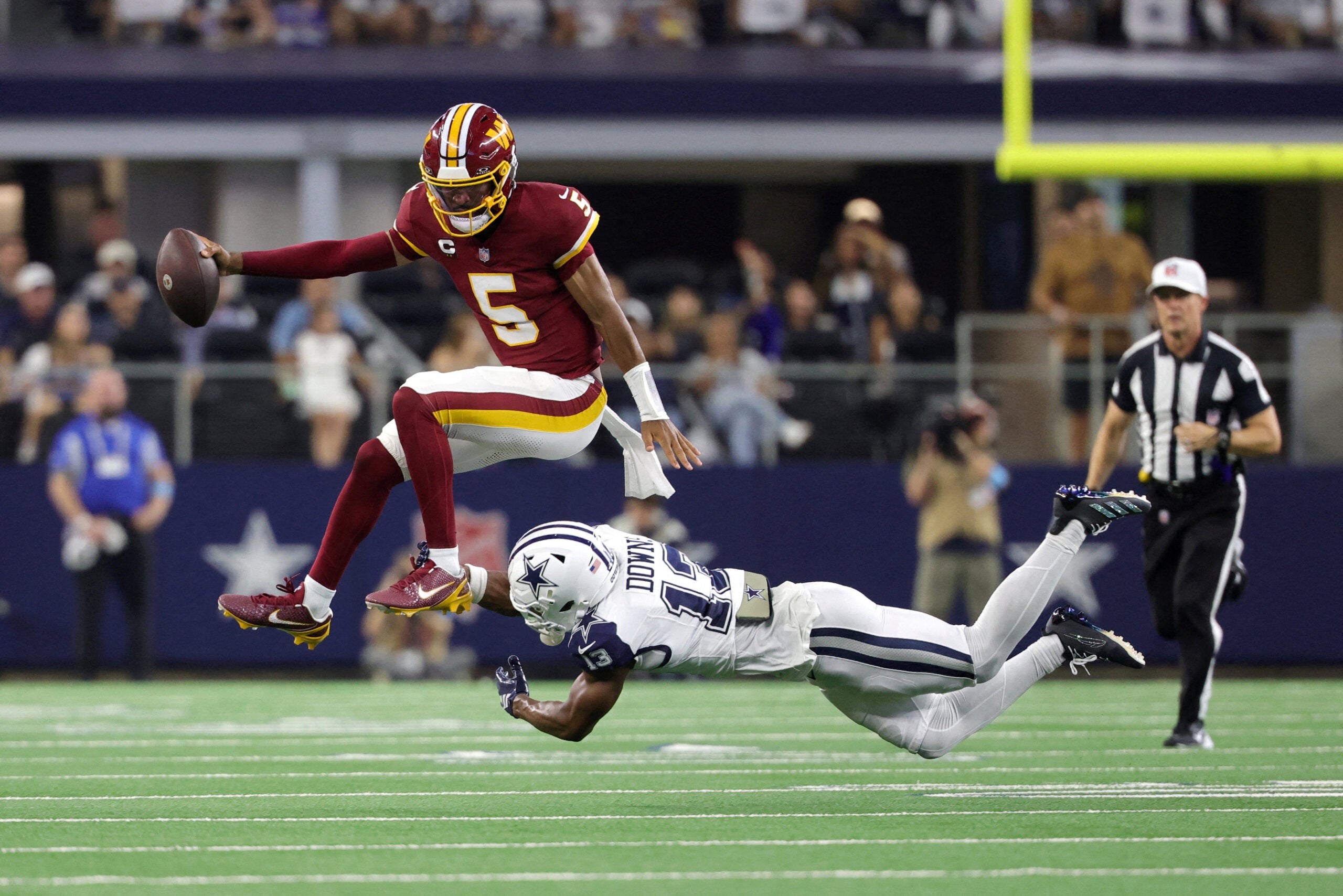 Sep 20, 2026; Arlington, Texas, USA; Washington Commanders quarterback Jayden Daniels (5) leaps to avoid a tackle by Dallas Cowboys safety Caleb Downs (13) while running with the ball during the second half at AT&T Stadium.